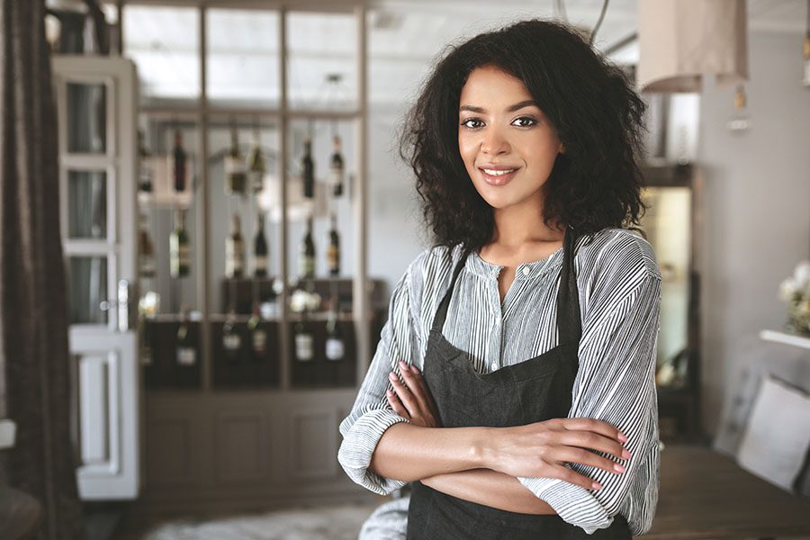 Business Insurance - Portrait of Woman Standing With Clasped Hands and Wearing an Apron in a Restaurant With Featured Wines Displayed and Blurred in the Background